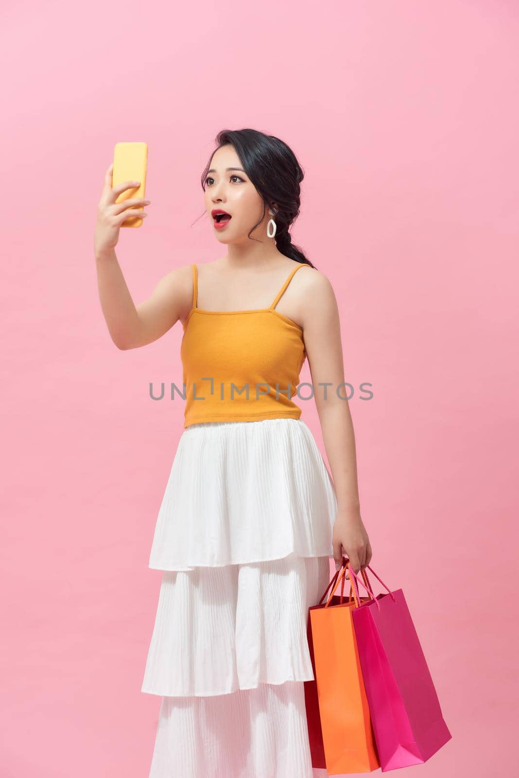 Surprised young woman with mobile phone and shopping bags on color background