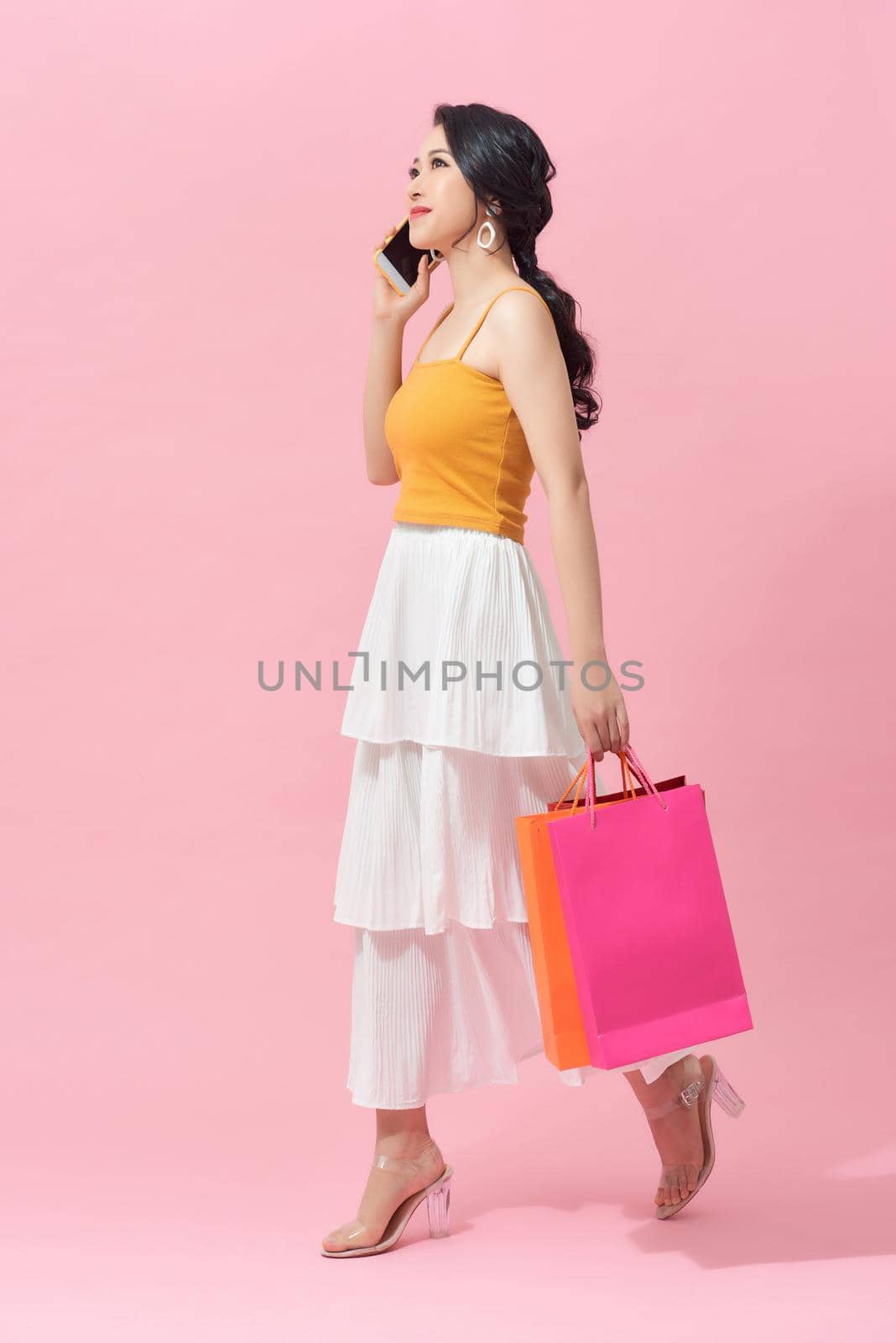 Full length of a beautiful young woman wearing colorful clothes standing isolated over pink background, carrying shopping bags, holding mobile phone by makidotvn