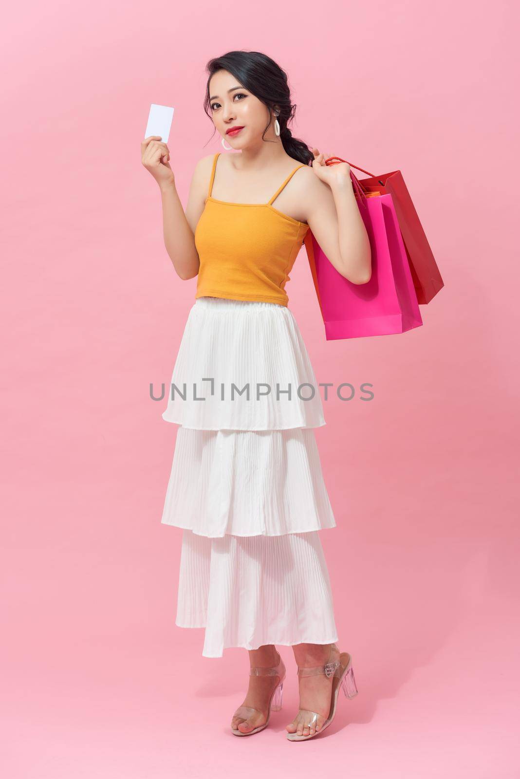 Full length of beautiful young Asian with colorful shopping bag and credit card over pink background. by makidotvn