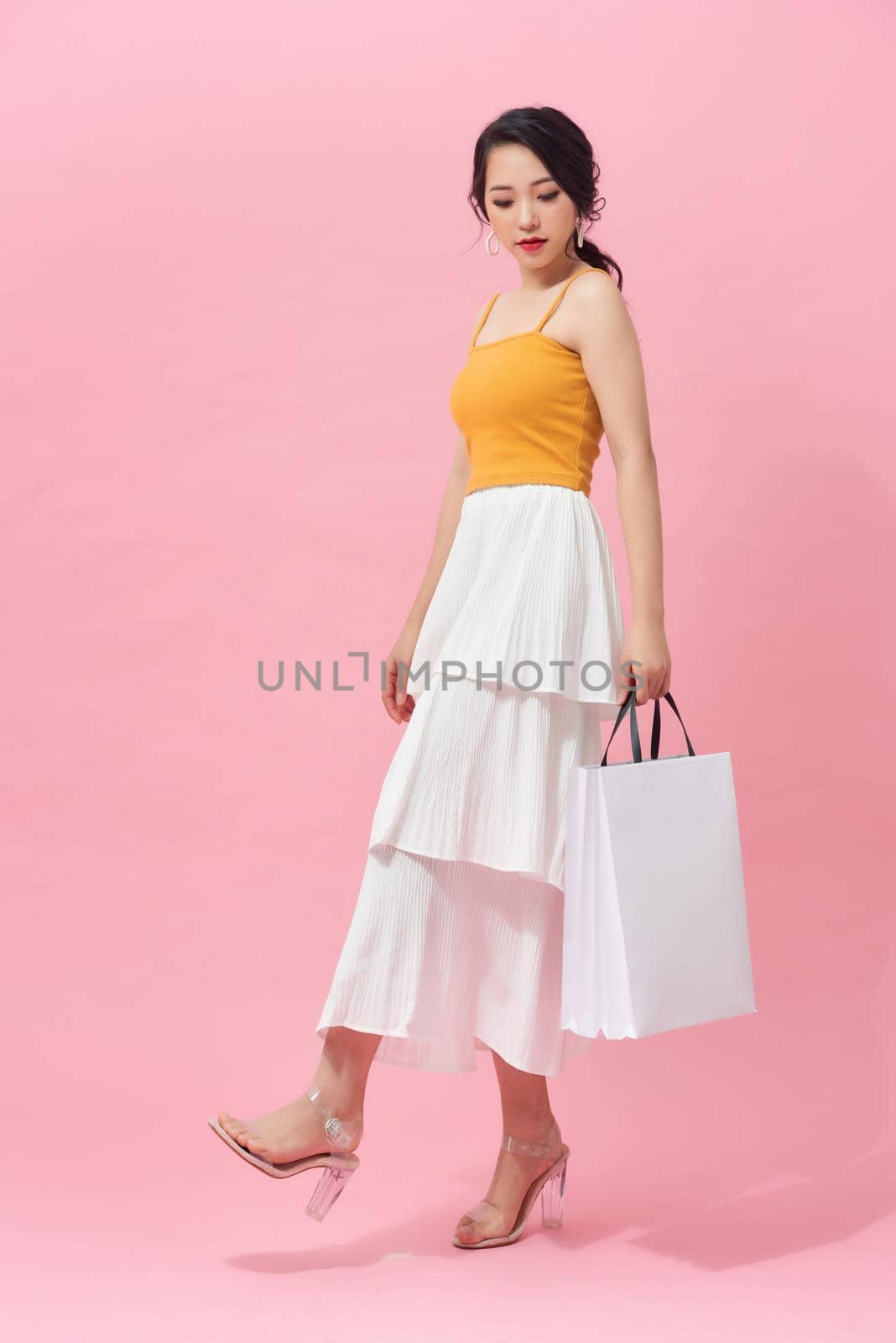 Full length side view of young woman walking with shopping bag isolated over pink background
