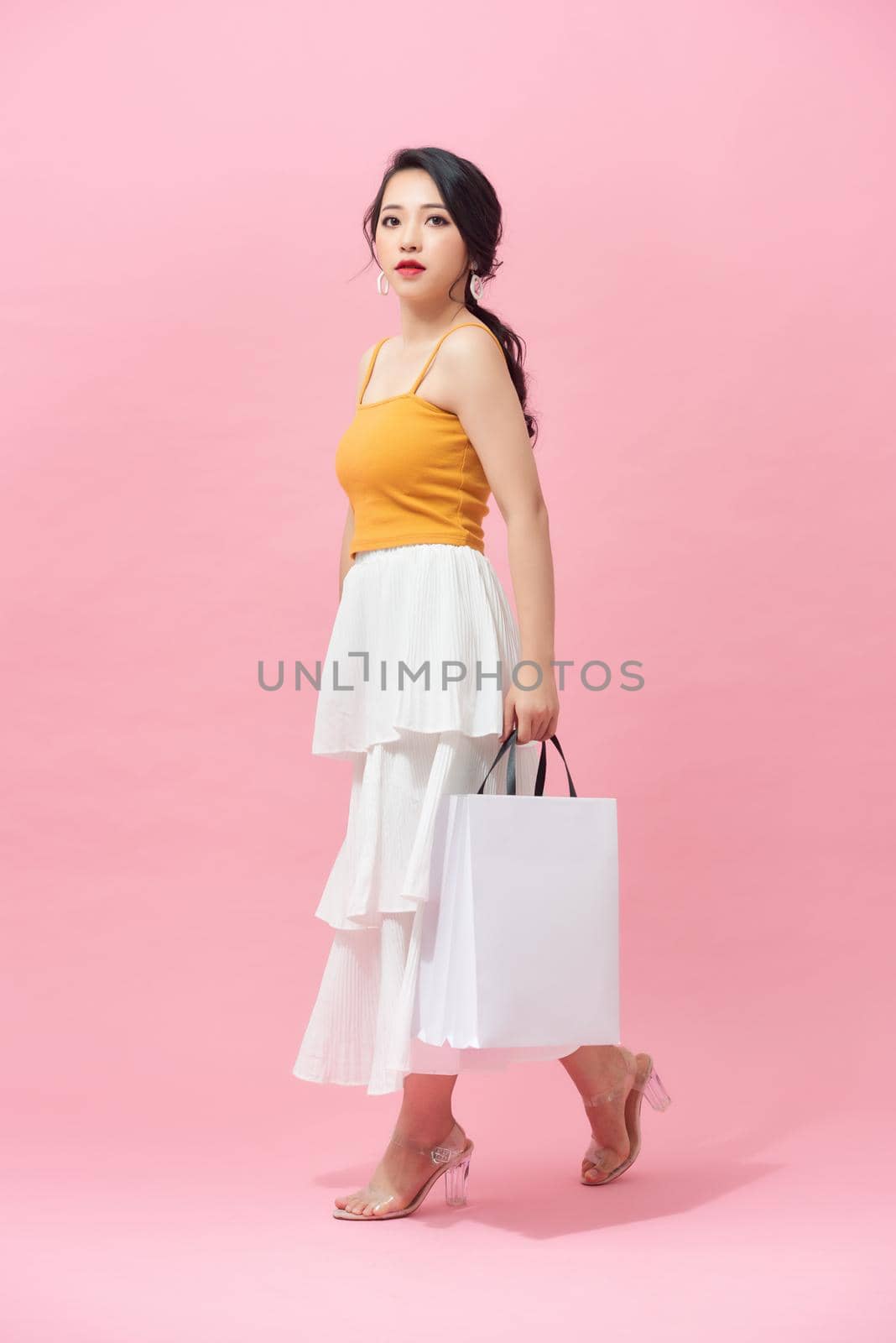 Full length of young woman holding shopping bags and smiling while walking isolated over pink background.