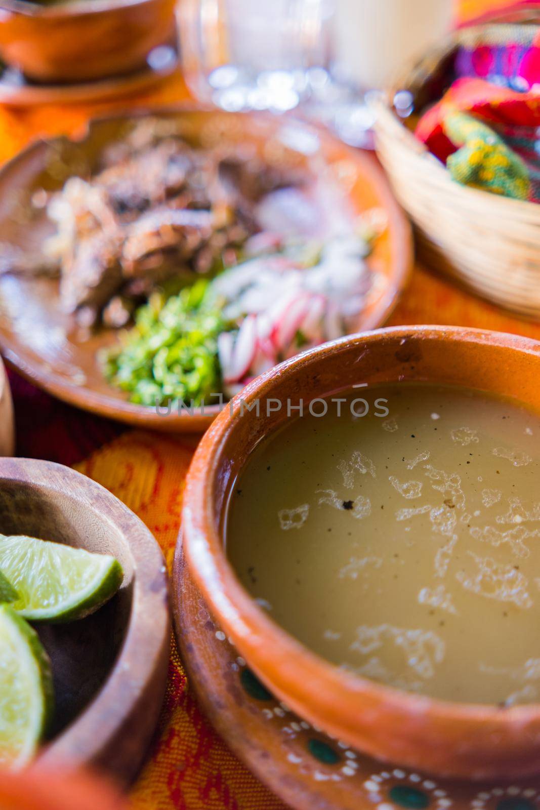 Close-up of clay bowl of lamb broth and plate of chopped lamb meat above orange and red tablecloth. Traditional soupy dish and condiments above colorful table. Classic Mexican cuisine