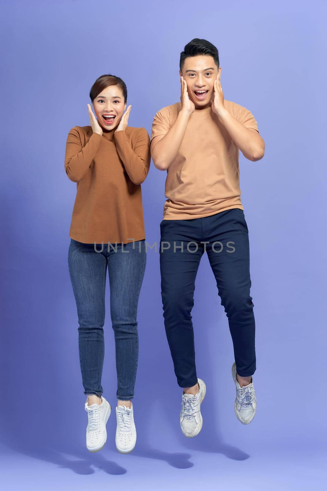 Happy couple shouting and touching cheeks while jumping with spreading legs isolated over purple background