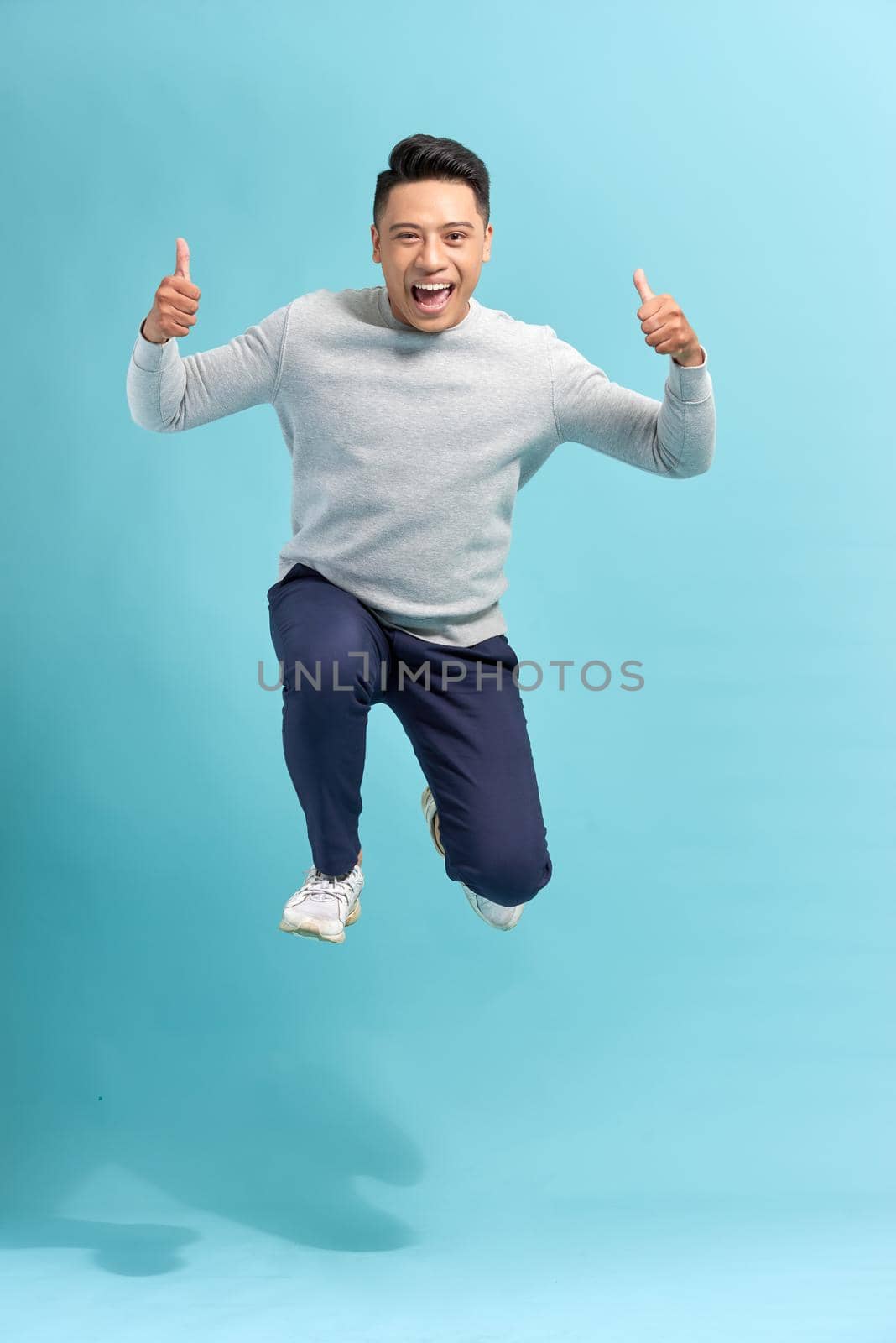 Full length portrait of an excited bearded man jumping and showing thumbs up isolated over blue background