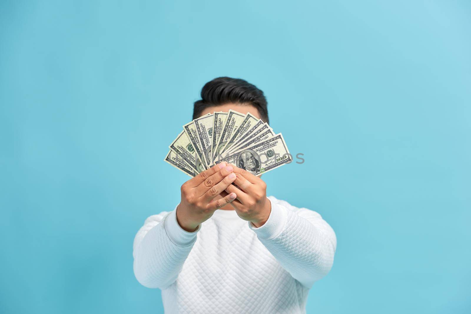 Photo of funny man in casual t-shirt covering his face with fan of money in dollar currency isolated over blue wall