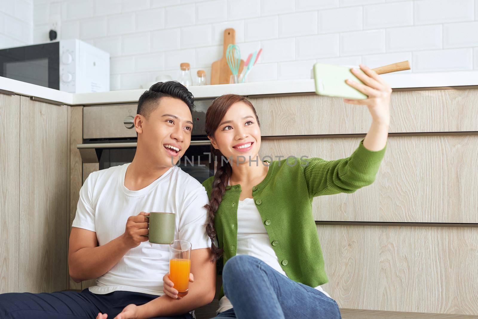 Beautiful young couple in love sitting on the kitchen floor, taking selfies using smart phone, by makidotvn