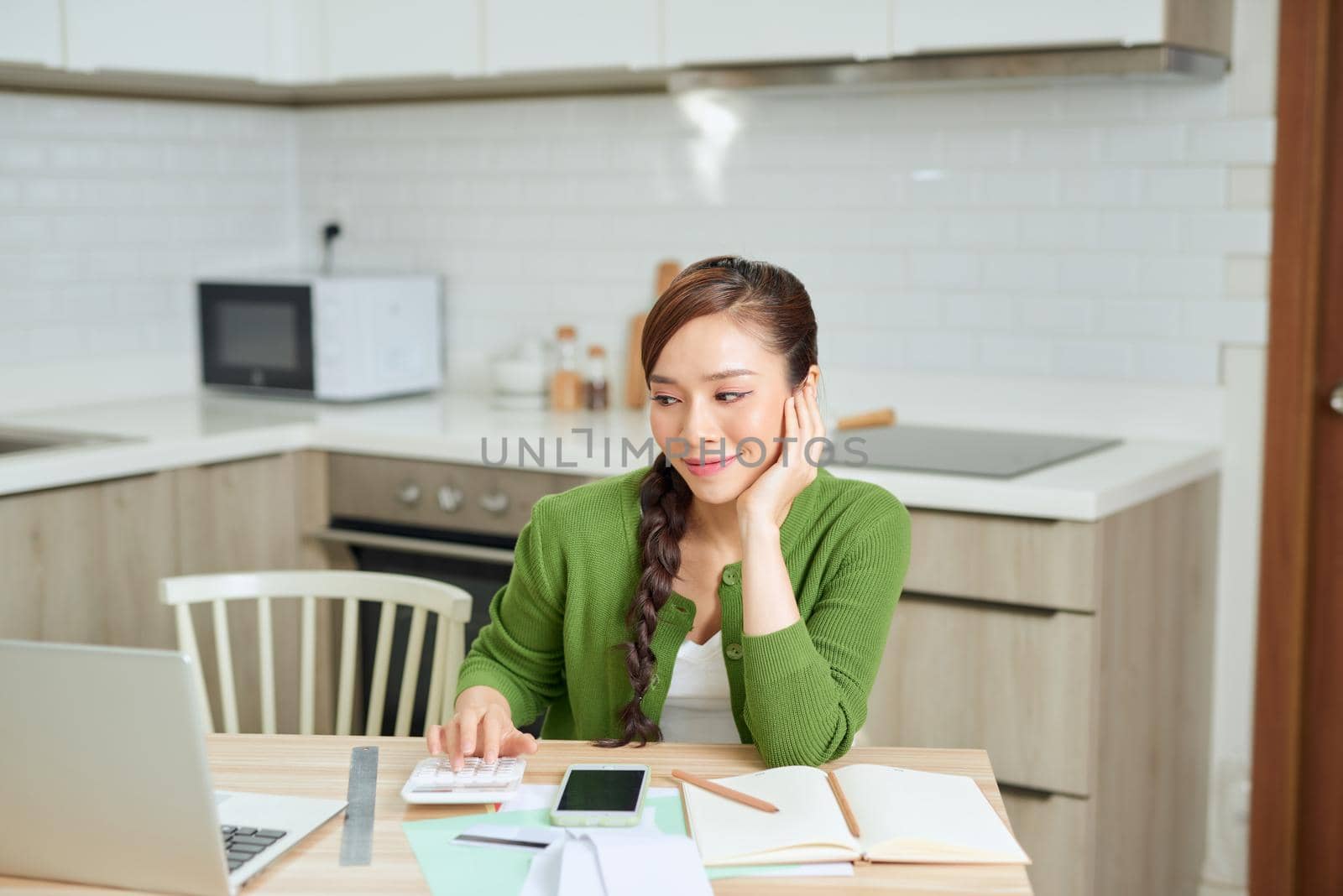 Attractive young smiling Woman paying bills online using laptop from home while sitting in the kitchen