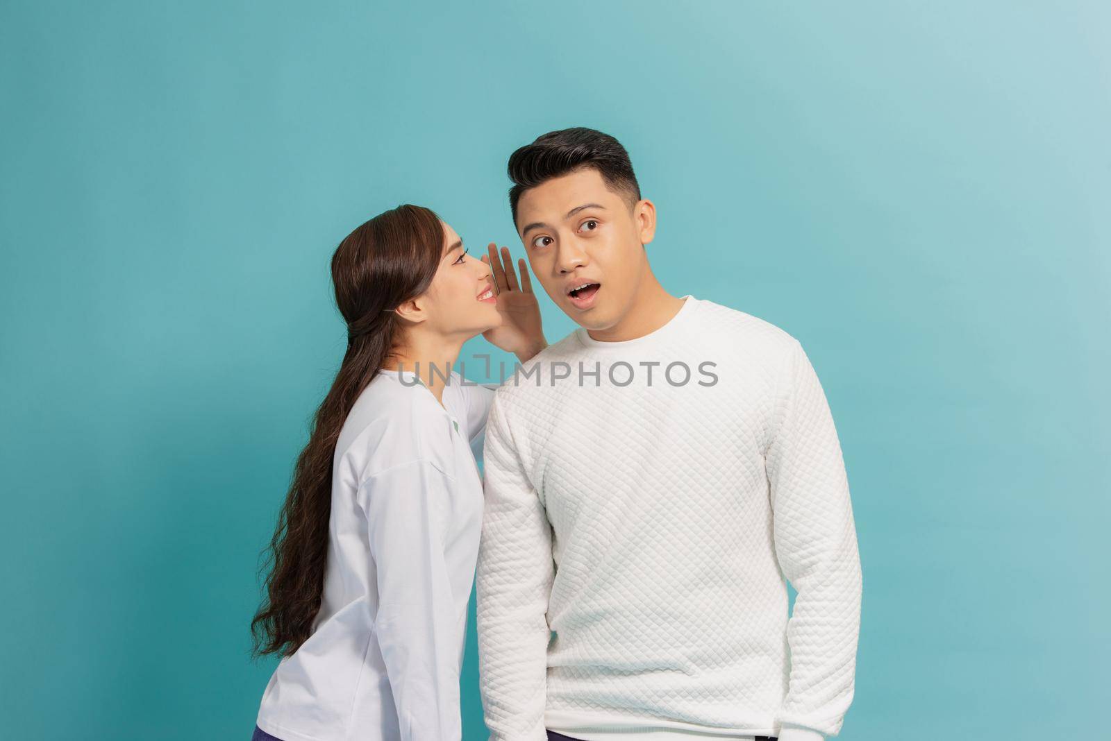 Portrait of a happy young couple standing together over blue background, telling secrets