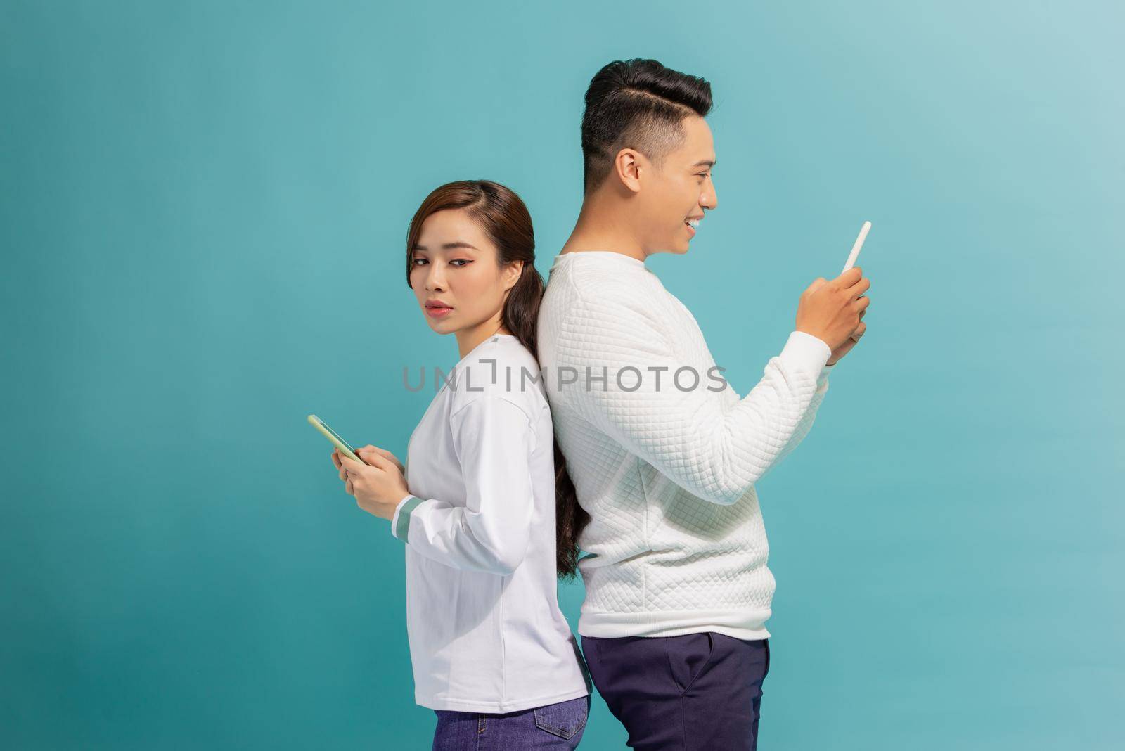 Asian girl and smile guy standing back to back, using mobile phones over blue background