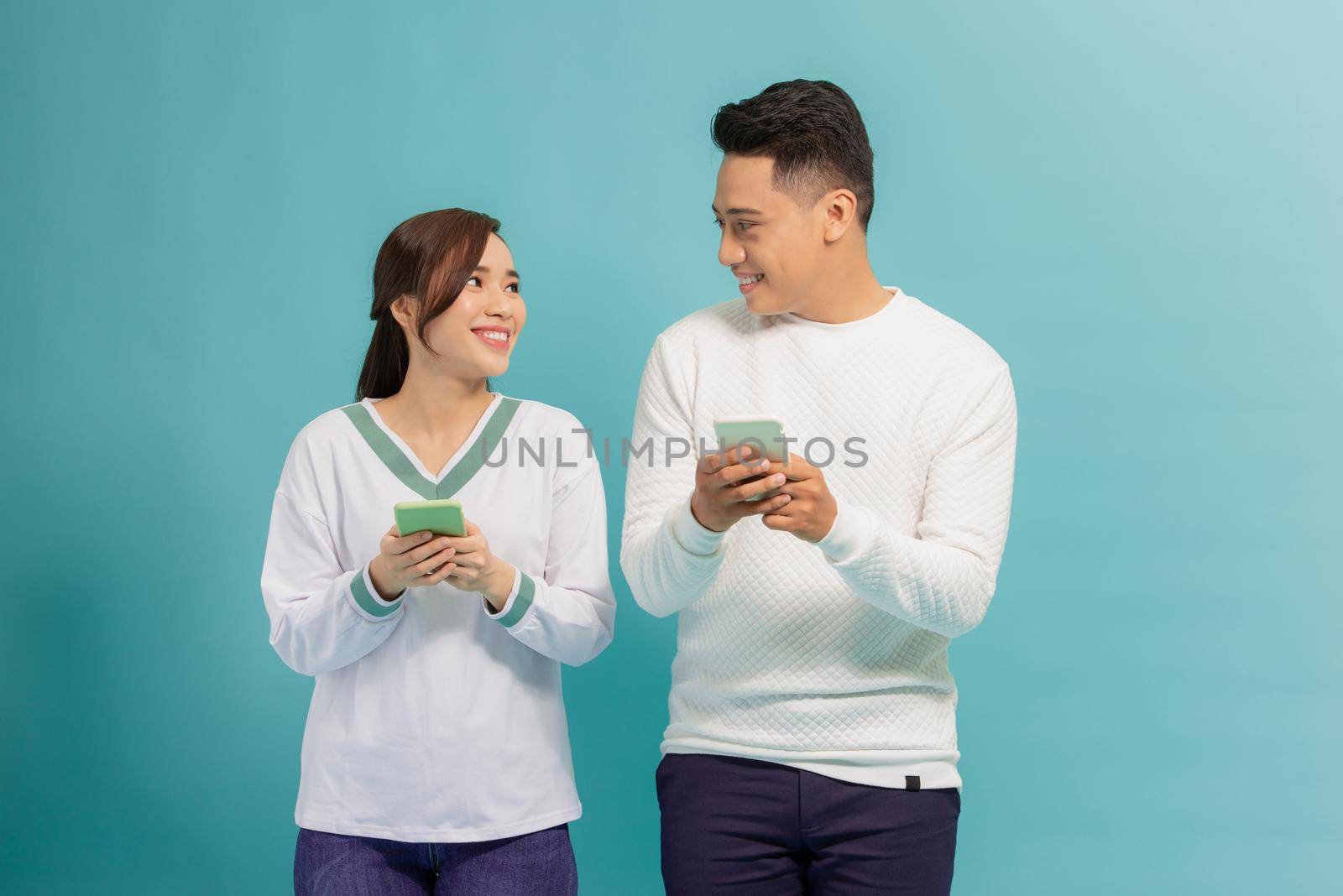 Cheerful young couple standing isolated over blue background, holding smart phones in casual wear by makidotvn