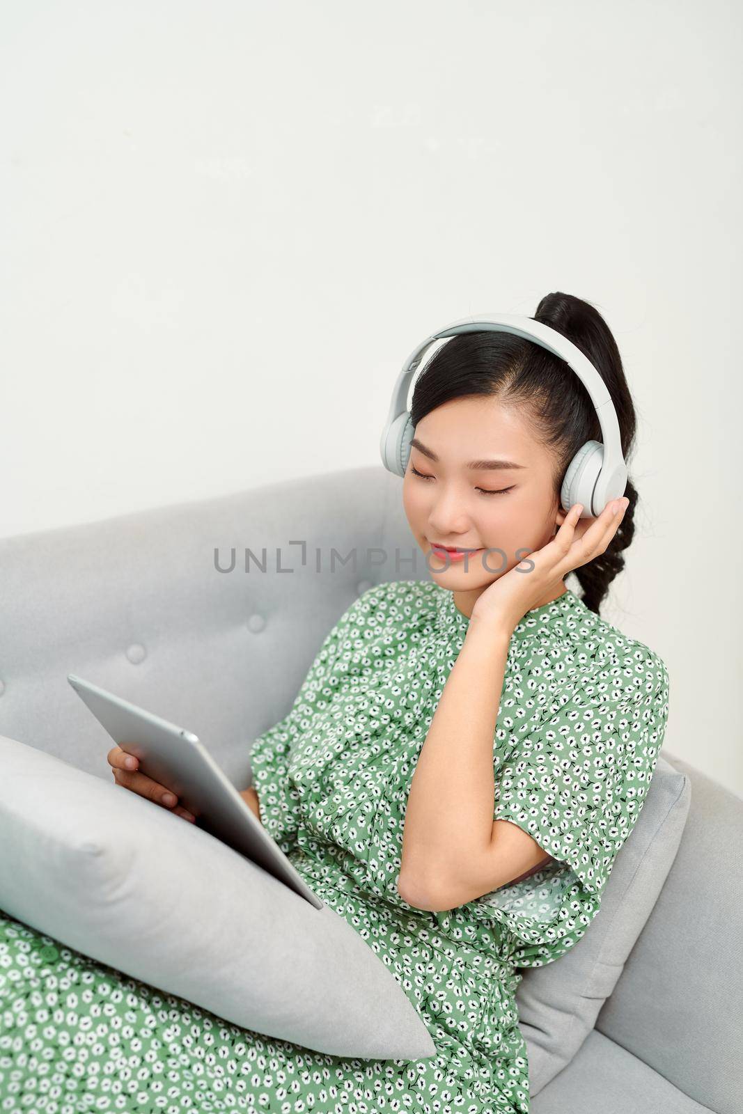 Young beautiful Asian woman laying in sofa and playing tablet with headphones 