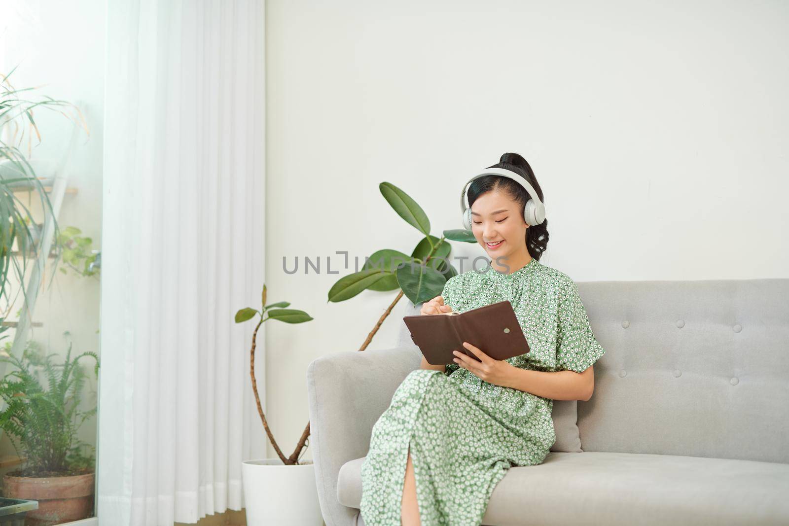 Young Asian woman listening music from headphones and writing note for her work idea in diary book.