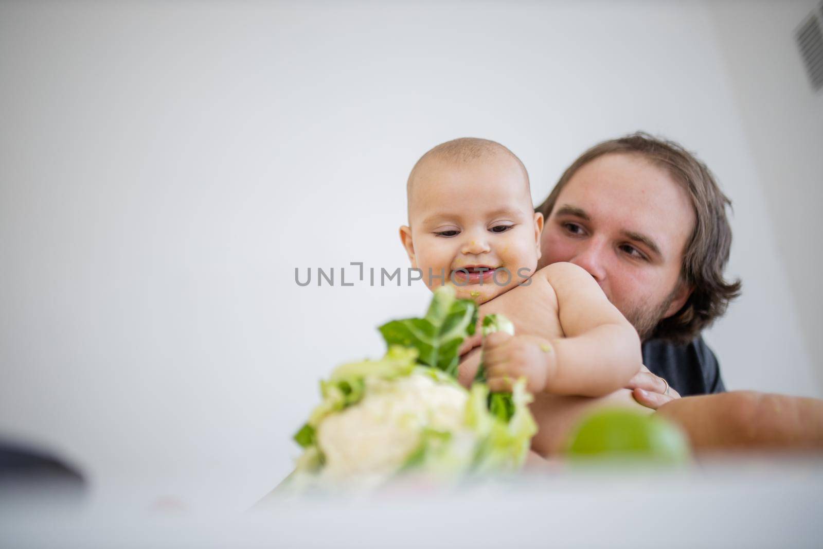 Father lovingly holding and kissing his happy baby daughter above table. Adorable baby smiling and playing with cauliflower. Babies interacting with food