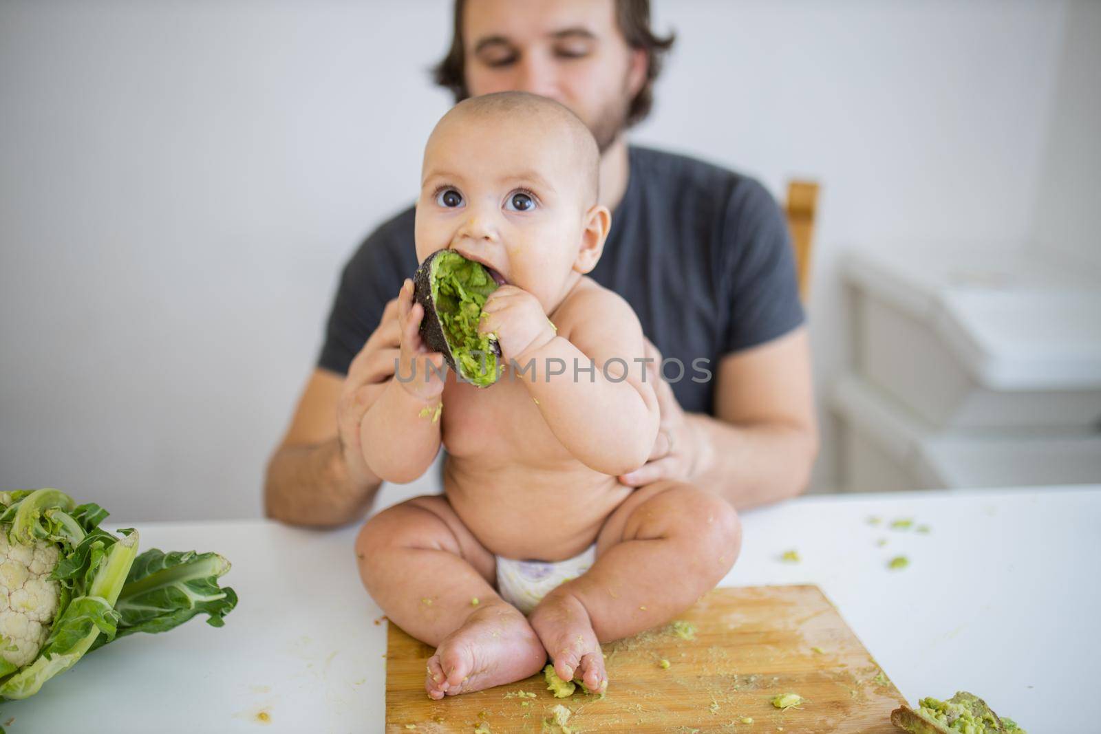 Father lovingly holding his happy baby daughter above table. Adorable baby sitting on wooden board and biting avocado peel. Babies interacting with food