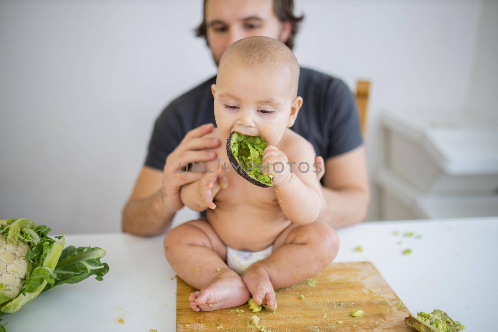 Father lovingly holding his happy baby daughter above table. Adorable baby sitting on wooden board and biting avocado peel. Babies interacting with food
