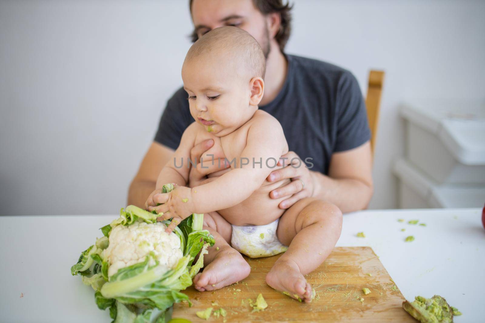 Father lovingly holding his happy baby daughter above table. Adorable baby on wooden board playing with cauliflower. Babies interacting with food