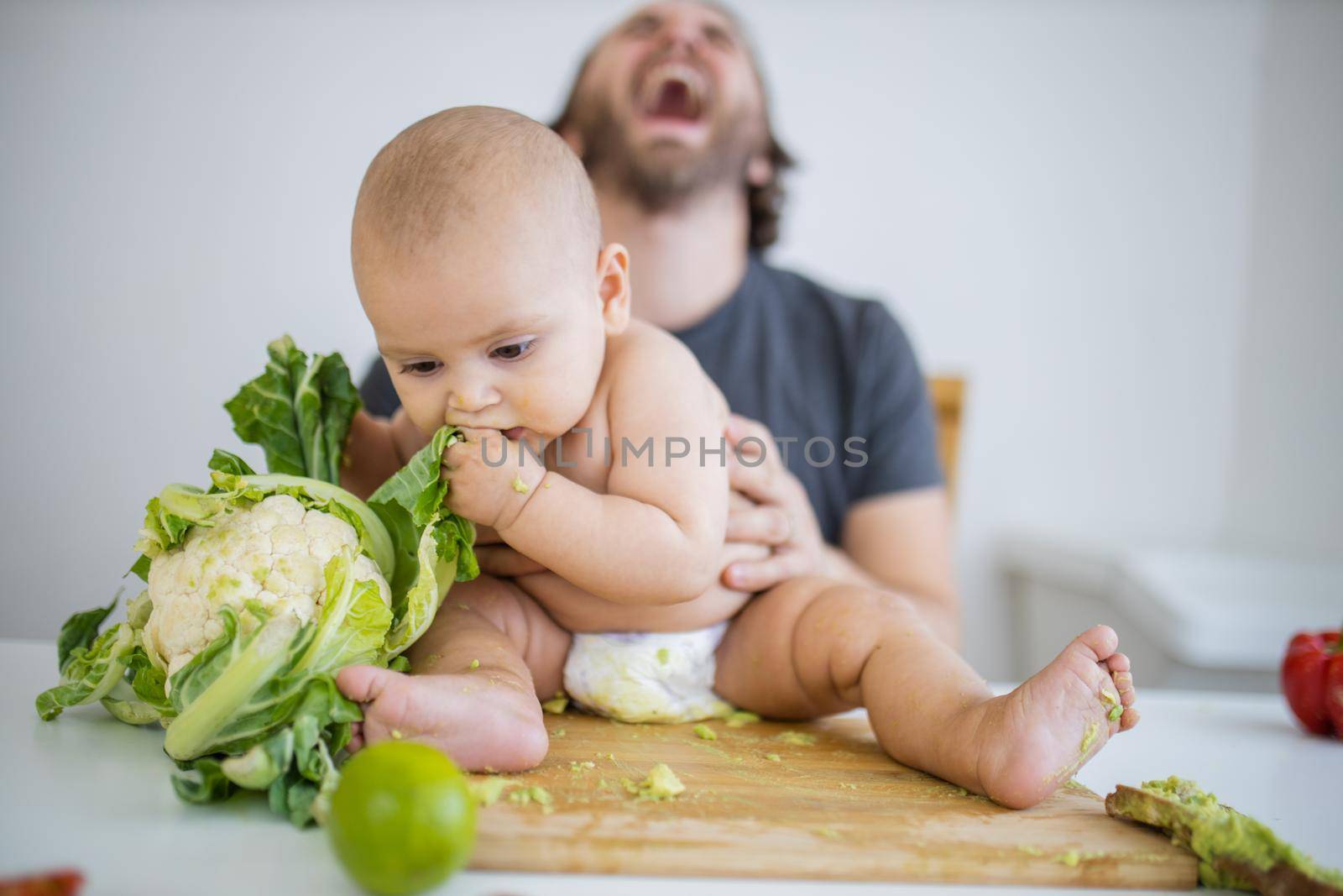 Father laughing hysterically and holding his happy baby daughter above table. Adorable baby sitting on wooden board and biting cauliflower. Babies interacting with food