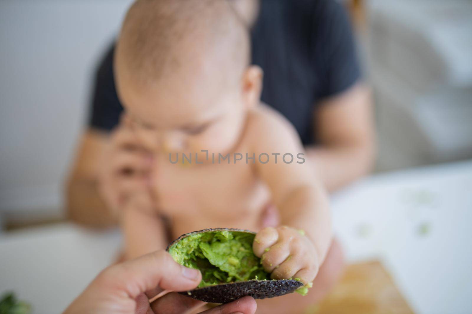 Adorable baby sitting on white table and holding an avocado peel. Father lovingly holding his happy daughter above wooden table. Babies interacting with food