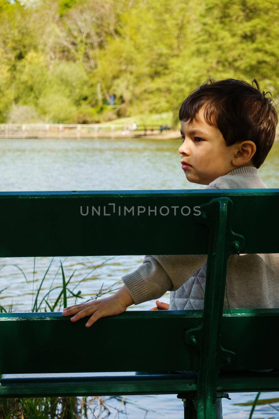 Little boy sitting on a bench next to a lake, spring season