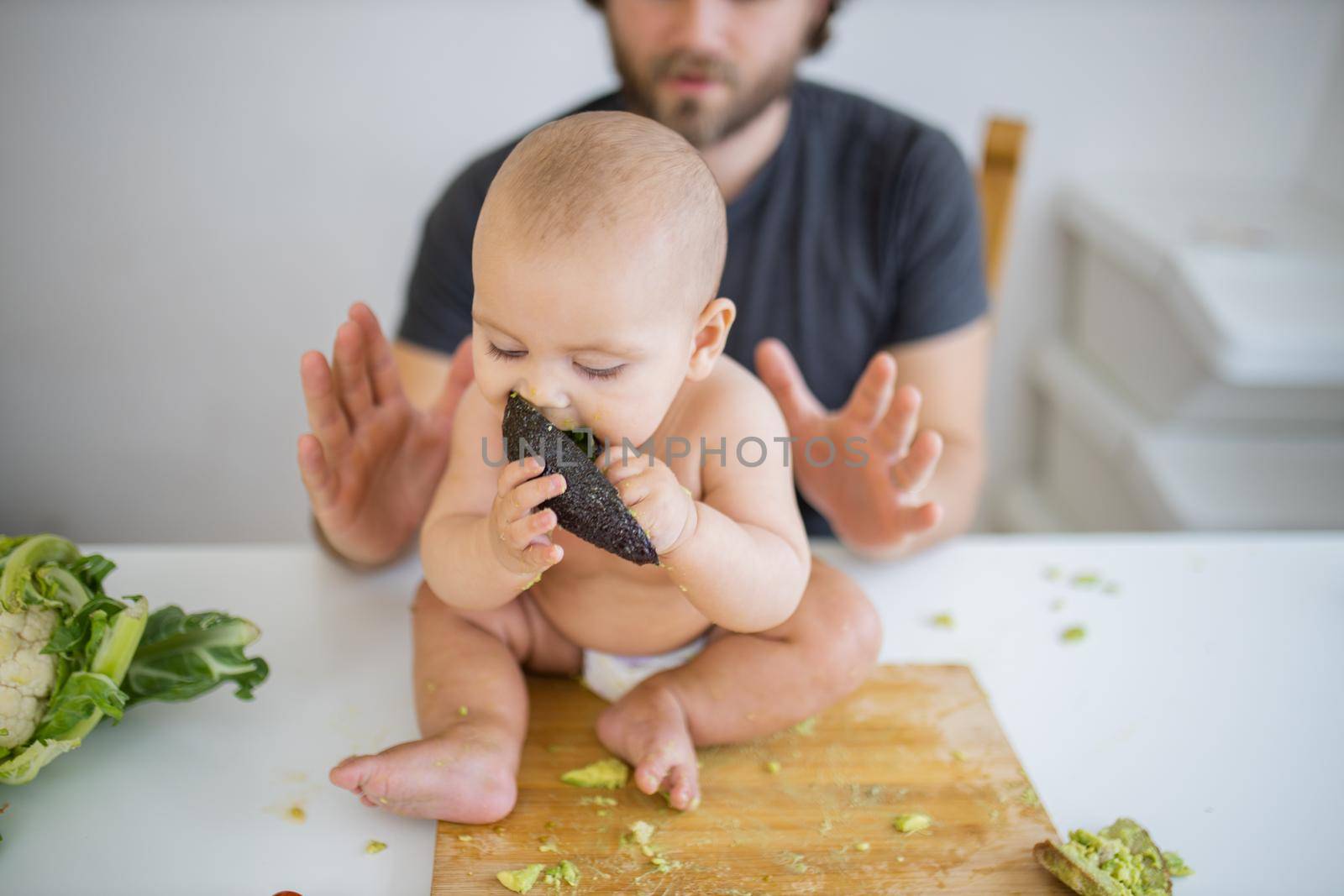 Happy baby sitting on wooden board and adorably biting avocado peel. Father lovingly holding his happy baby daughter above table. Babies interacting with food