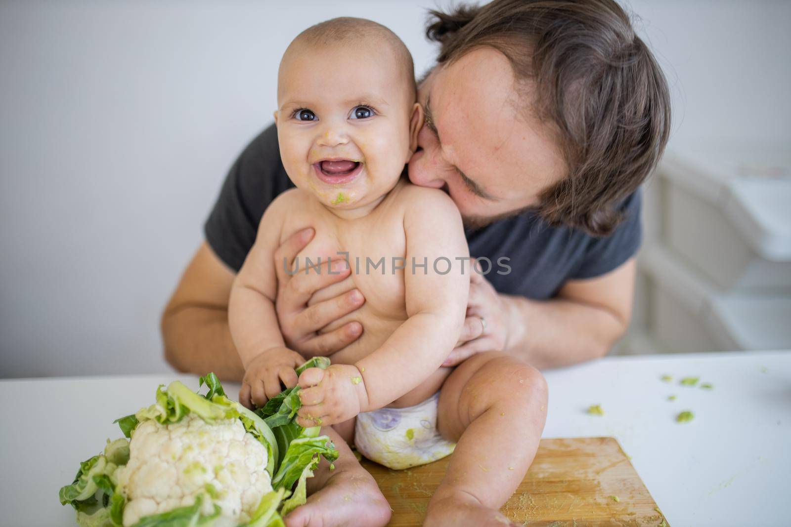 Father lovingly holding and kissing his happy baby daughter above table. Adorable baby on wooden board smiling and holding cauliflower. Babies interacting with food