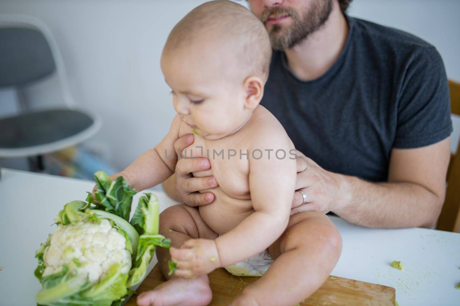 Father lovingly holding his happy baby daughter above table. Adorable baby on wooden board playing with cauliflower. Babies interacting with food