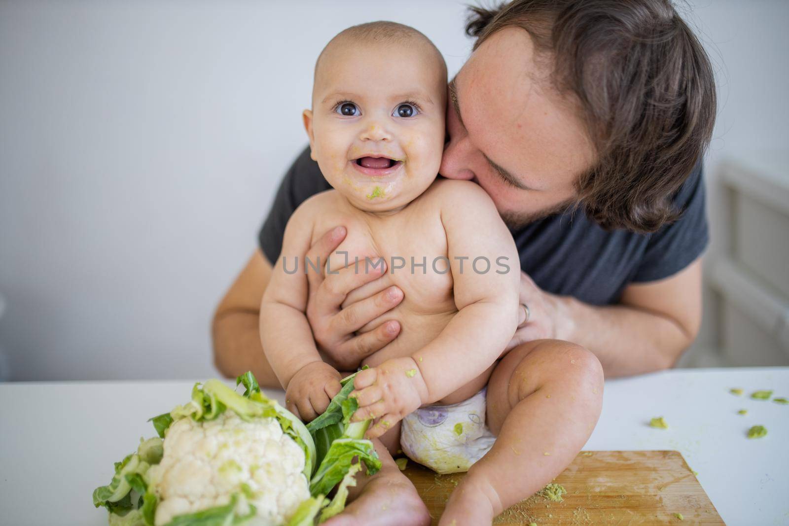 Father lovingly holding and kissing his happy baby daughter above table. Adorable baby on wooden board smiling and holding cauliflower. Babies interacting with food