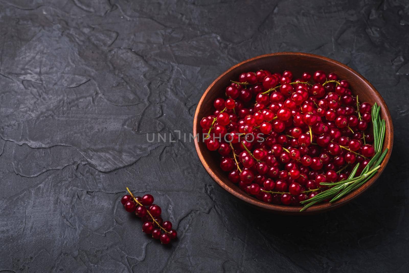 Fresh sweet red currant berries with rosemary leaves in wooden bowl, dark textured background, angle view copy space