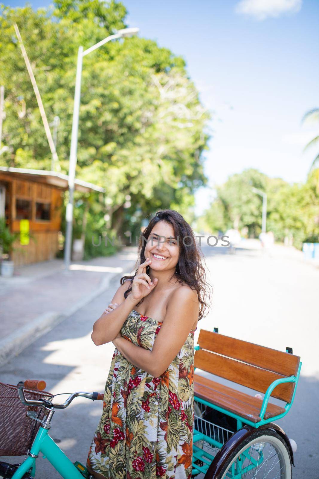 Beautiful smiling woman wearing flowered dress and riding adult tricycle with trees as background. Portrait of woman riding bike in empy street. Bike ride on sunny day