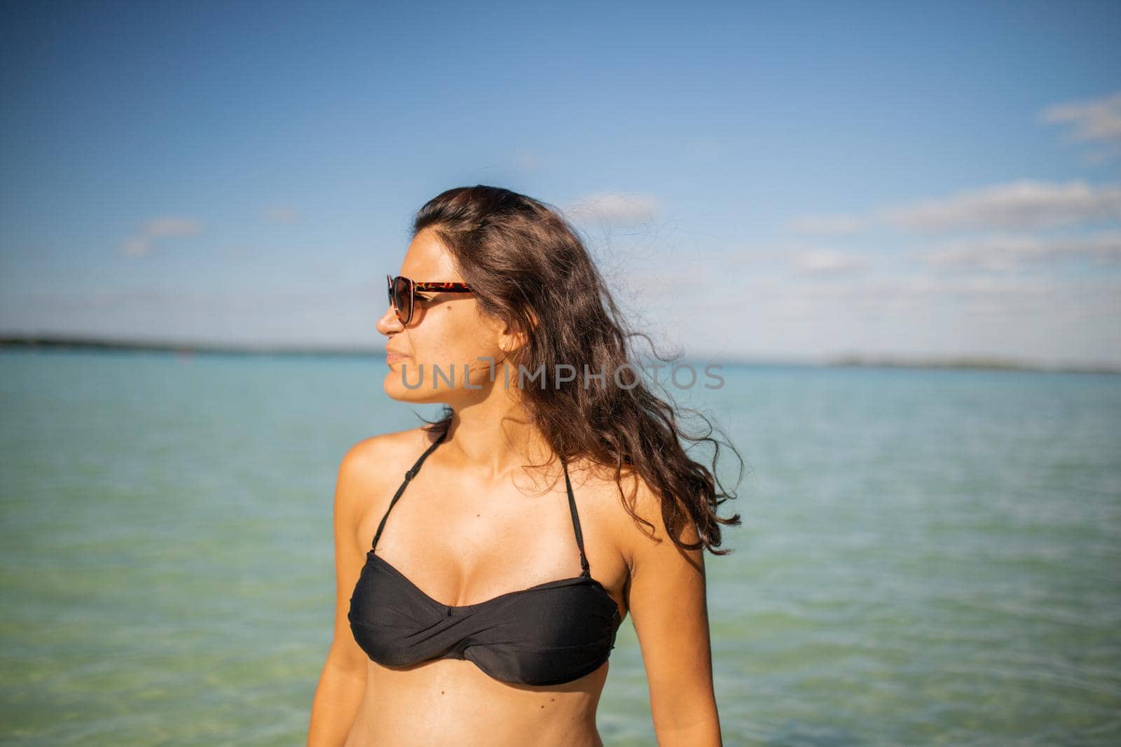 Beautiful brunette woman standing with blue sea and sky as background. Attractive woman wearing sunglasses and smiling serenely on beach. Tropical summer vacations