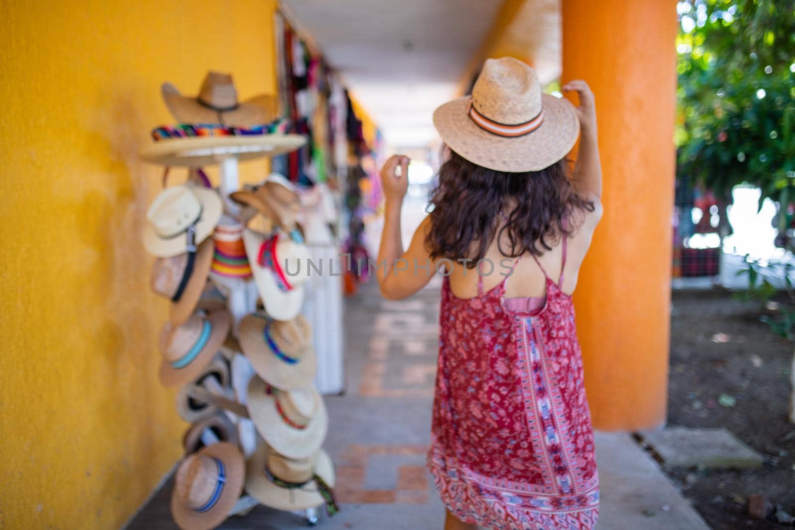 Brunette woman wearing red dress and straw hat standing next to hat rack. Young woman trying on different straw hats. Summer beauty and fashion