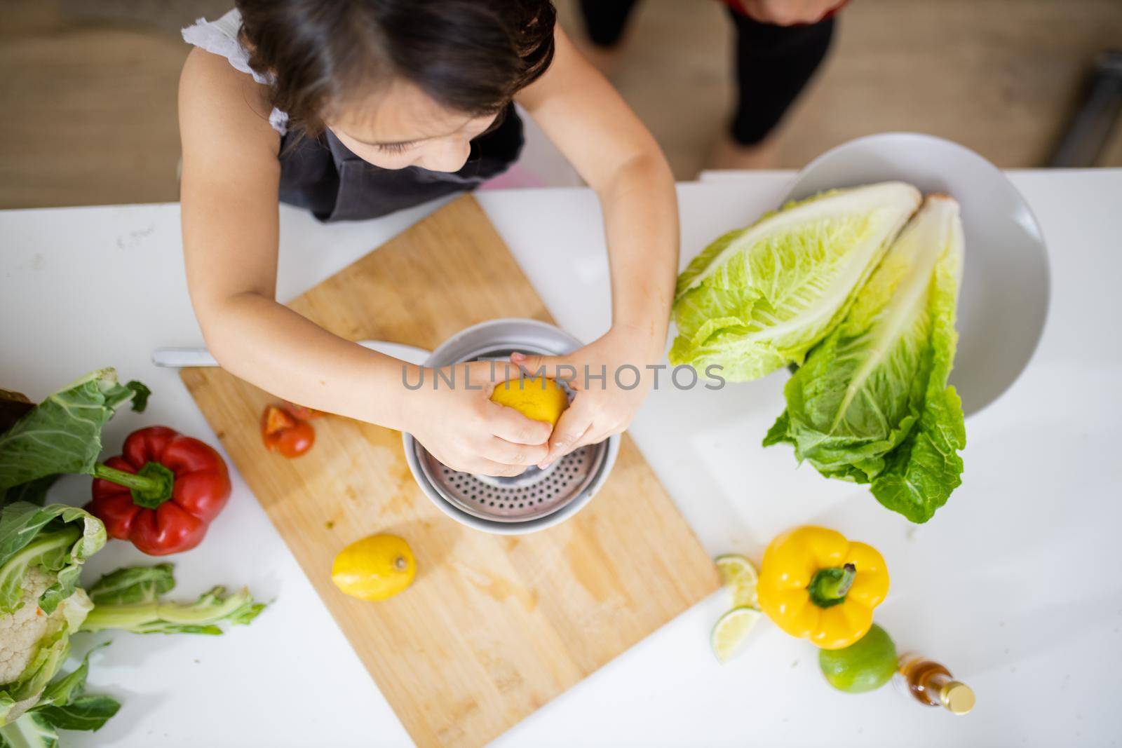 Adorable little girl squeezing lemon on steel lemon juicer surrounded by vegetables. Young child making juice on white table from above. Kids preparing food