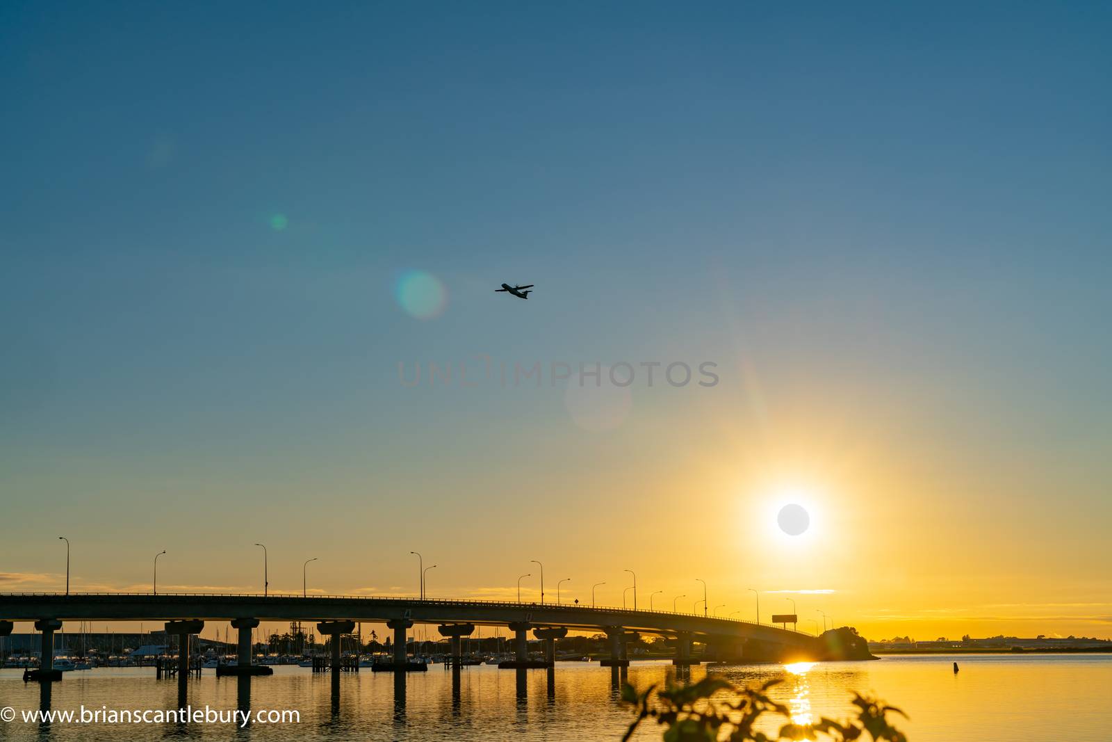 Sweeping lines of Tauranga Harbour Bridge over calm blue water with glow of rising sun at far end as passenger plane take-off and passes over