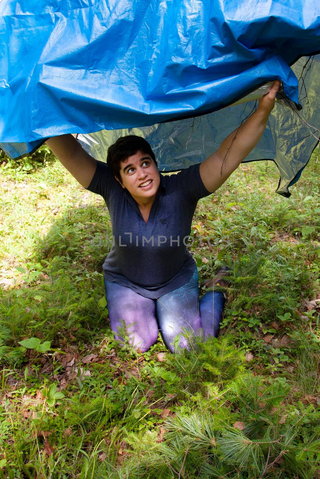 person takes shelter under a blue tarp in the forest while camping 
