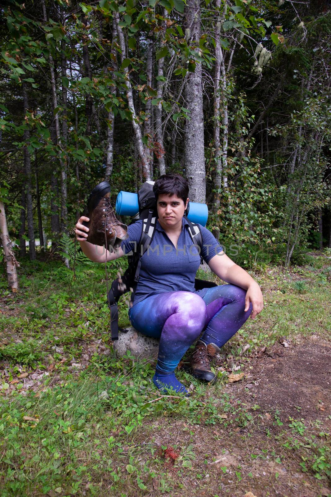 woman sitting on a rock pouring dirt and sand out of her hiking boot looking irritated 