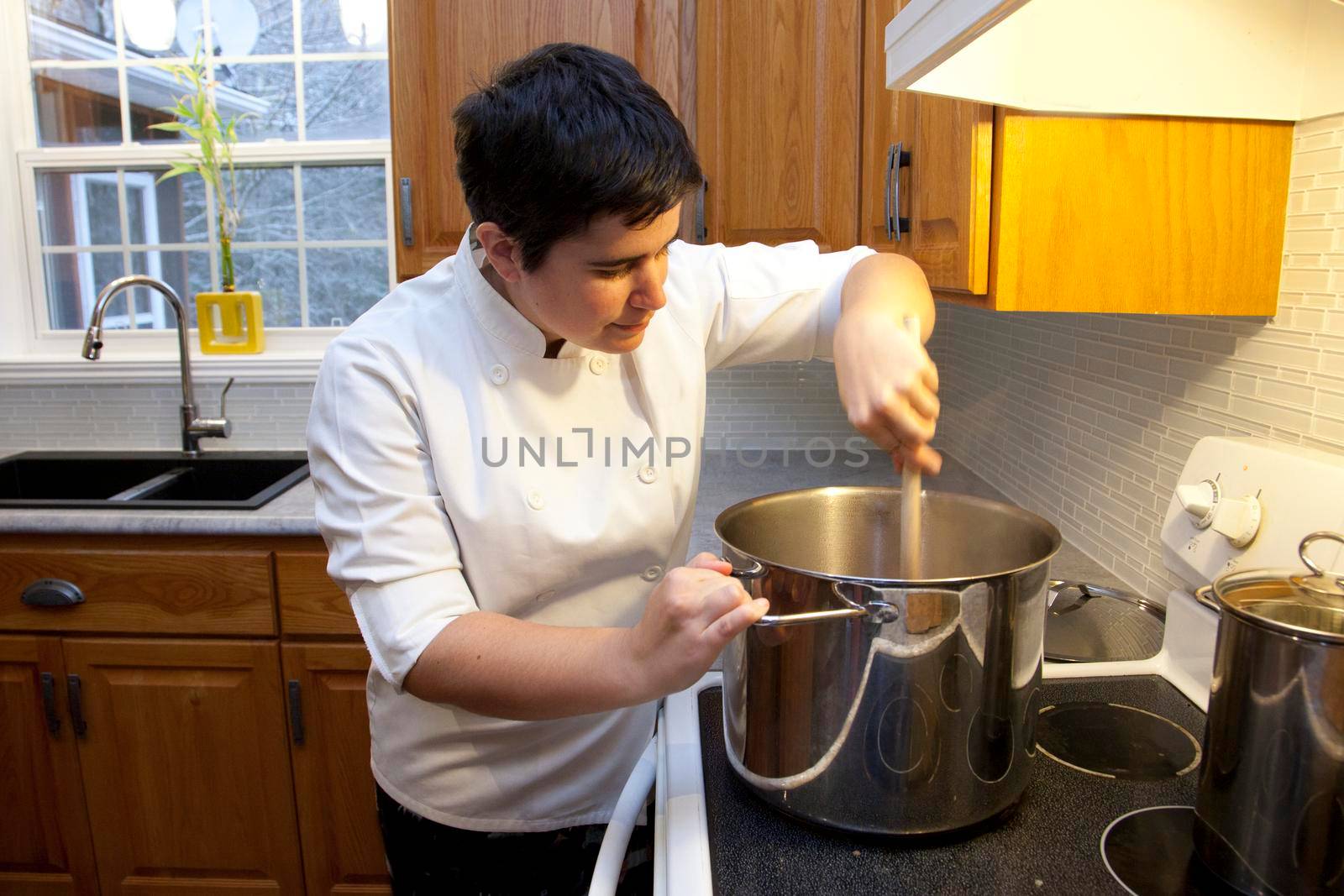  Person in white jacket looks into a huge vat while stirring with a wooden spoon in a beautiful kitchen