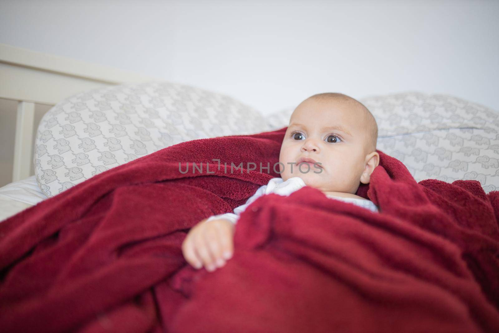 Relaxed-looking baby covered with a red blanket and lying on a bed by Kanelbulle