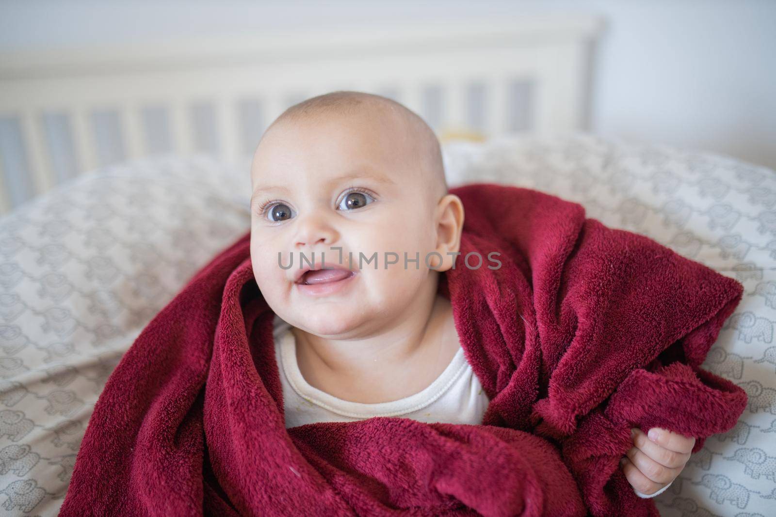 Adorable and happy baby covered with red blanket and lying on bed. Cute baby smiling and resting on pillows. Toddles and babies on beds