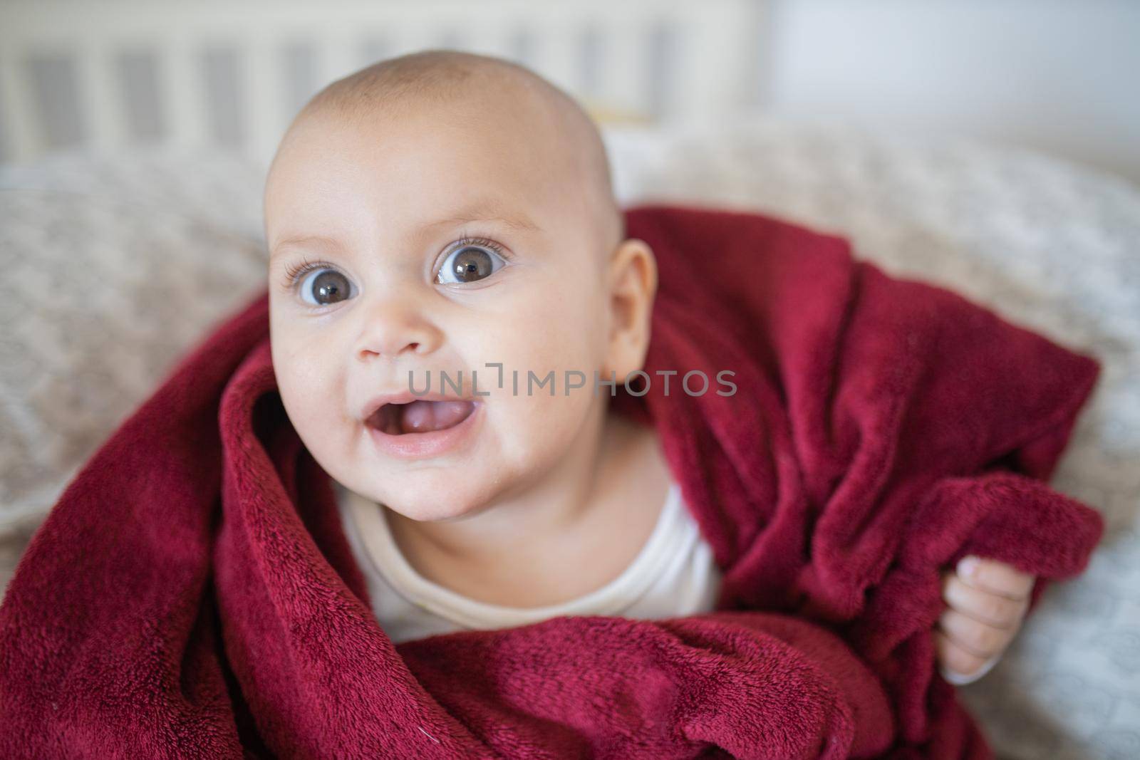 Adorable and happy baby covered with red blanket and lying on bed. Surprised-looking baby smiling and resting on pillows. Toddles and babies on beds