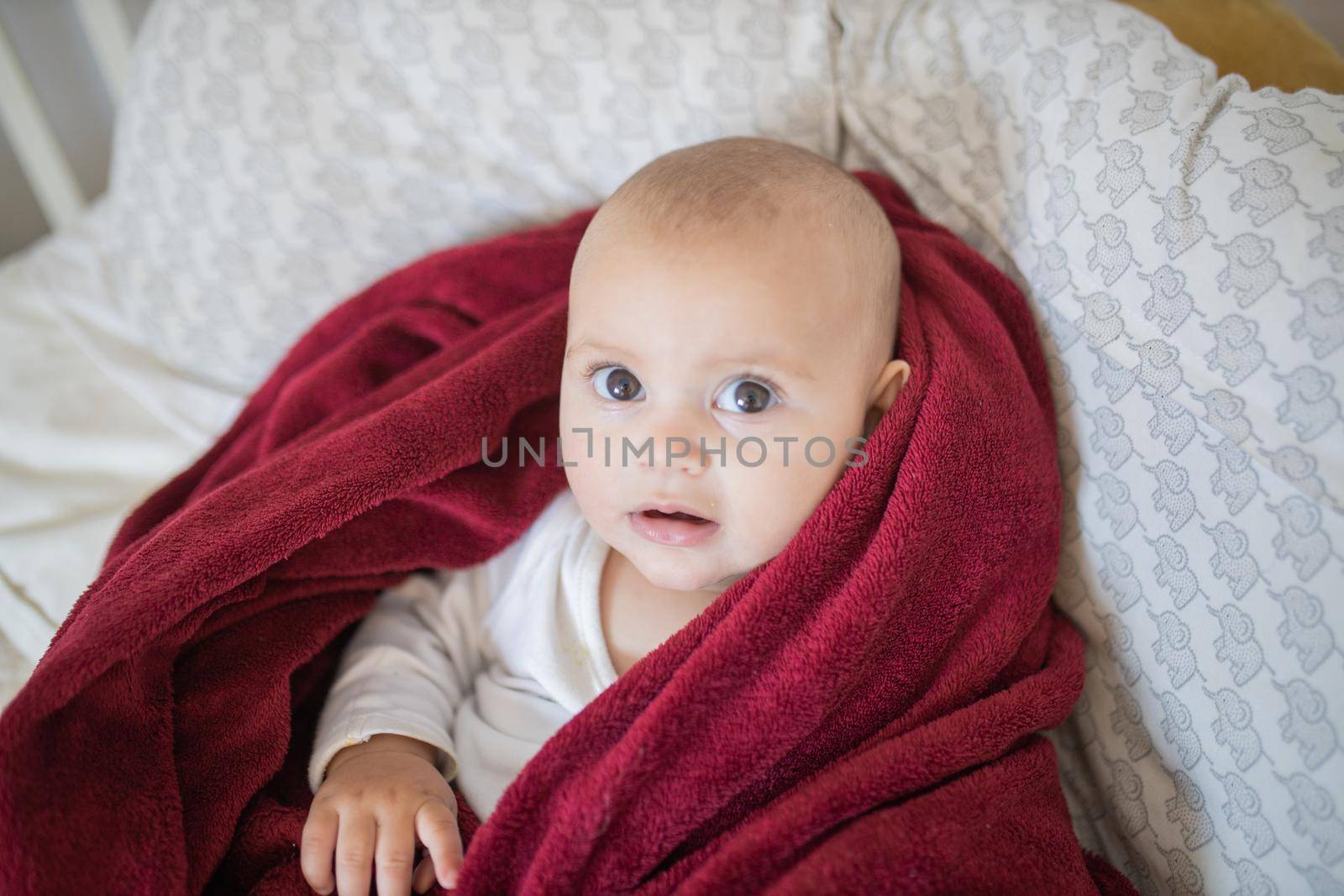 Concerned-looking baby covered with a red blanket and lying on a bed by Kanelbulle