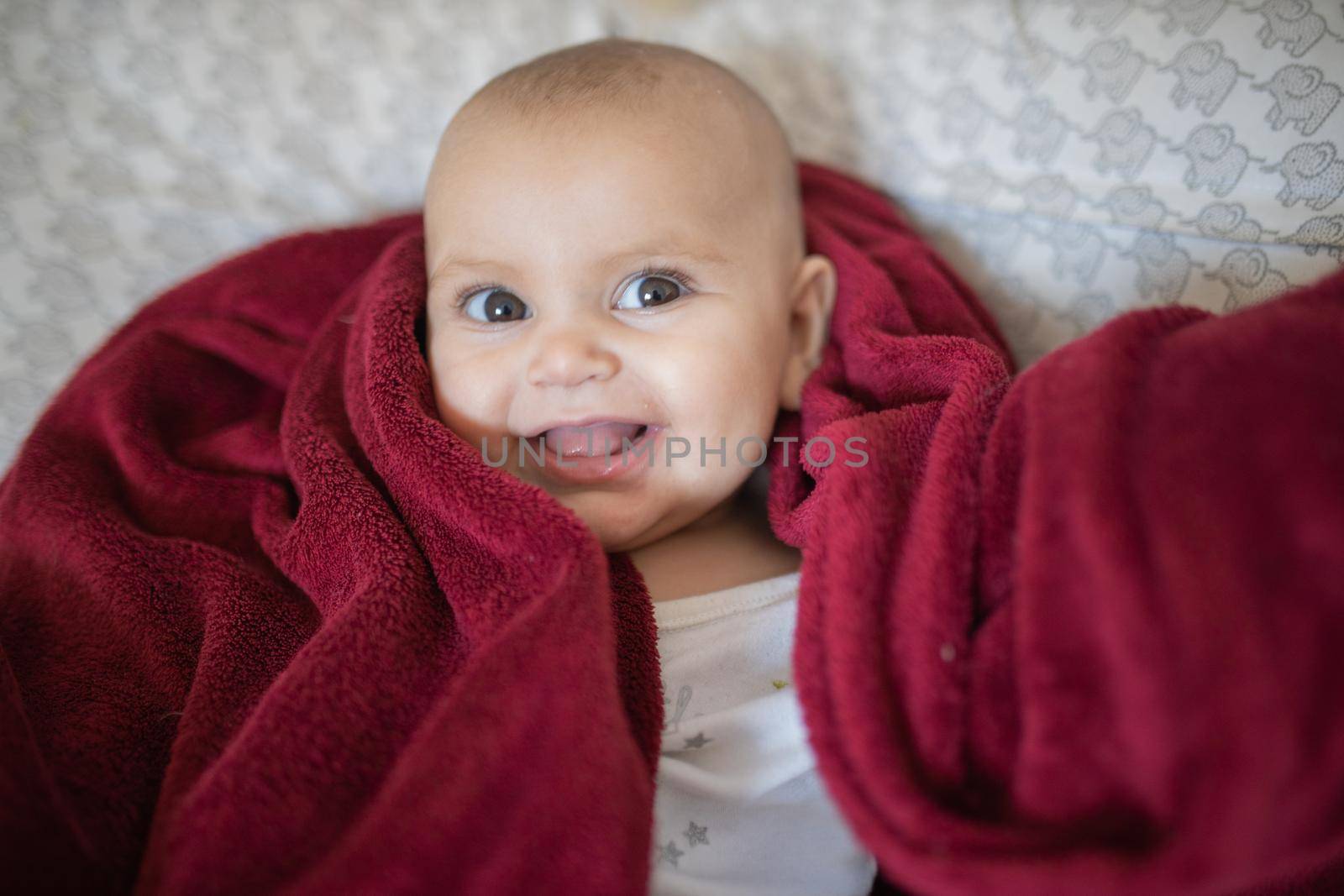 Adorable and happy baby covered with red blanket and lying on bed. Cute baby smiling and resting on pillows. Toddles and babies on beds