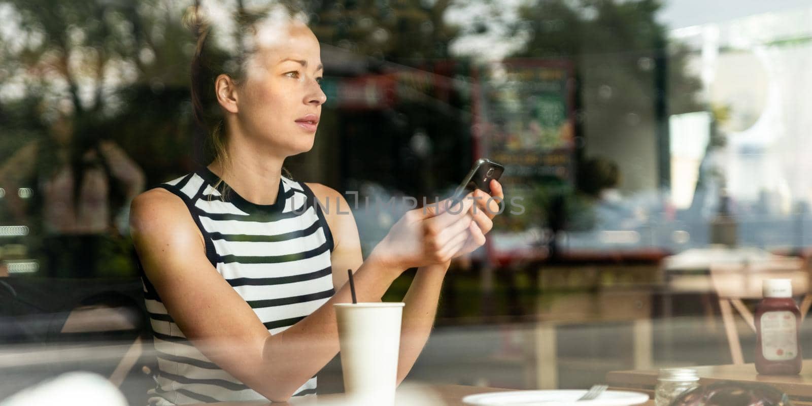Thoughtful casual caucasian woman holding mobile phone while looking through the coffee shop window during coffee break. Street reflections in the window glass.