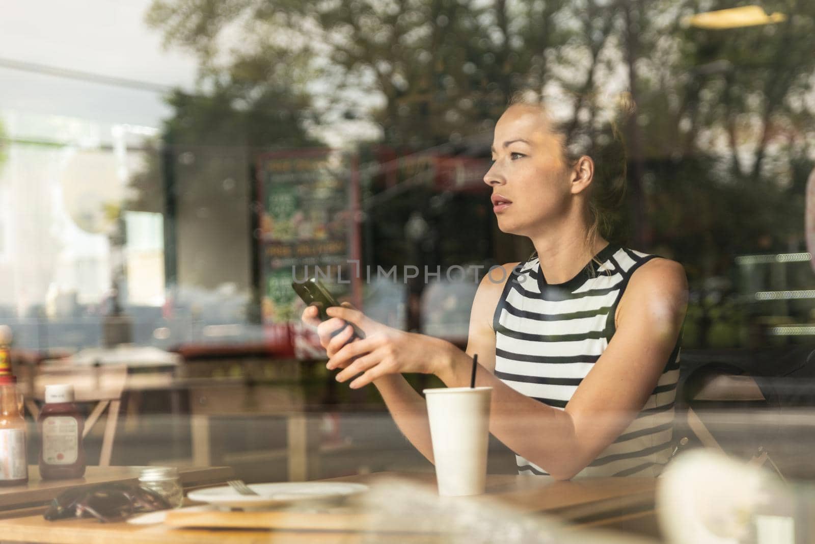 Thoughtful caucasian woman holding mobile phone while looking through the coffee shop window during coffee break. Street reflections in the window glass.