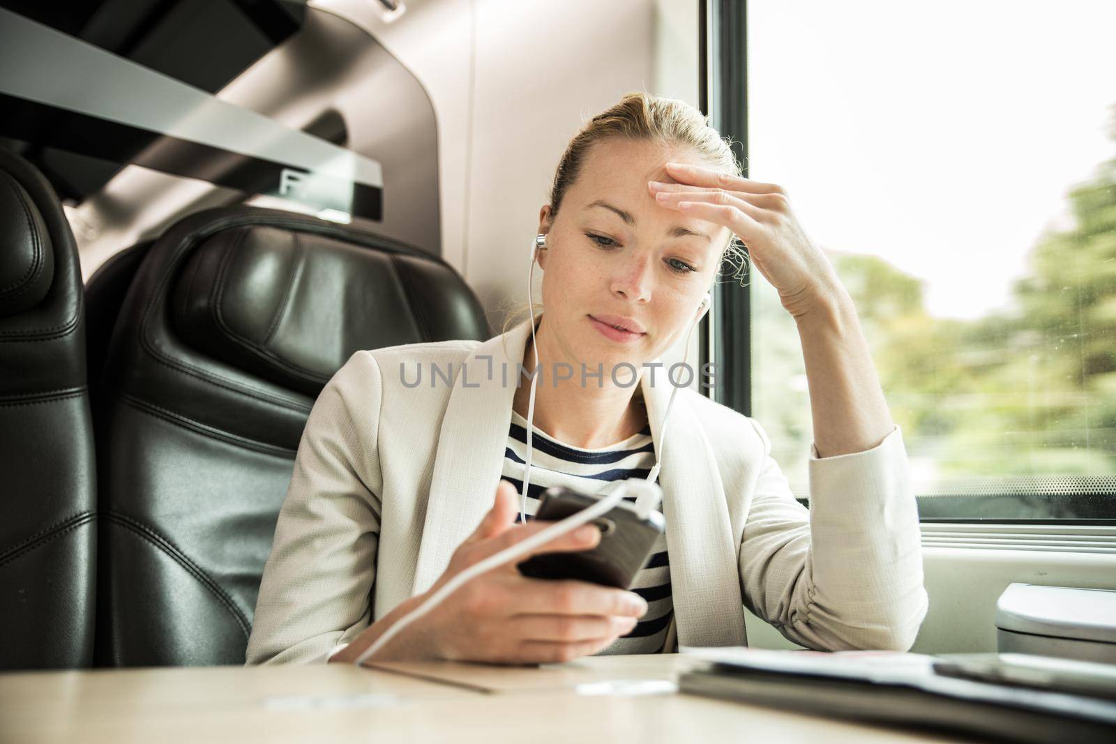 Businesswoman communicating on cellphone using headphone set while traveling by train in business class seat.