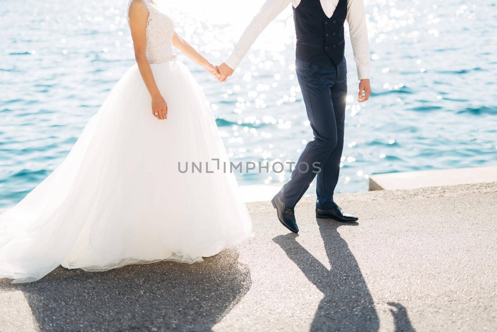 The bride and groom are walking hand in hand on the road by the sea, close-up. High quality photo