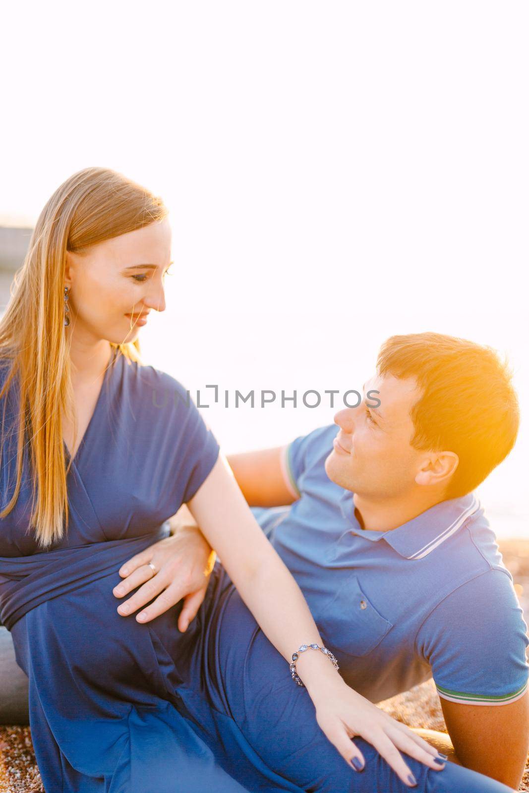 Man holds his hand on the belly of a smiling pregnant woman in a long blue dress while lying on the sand. High quality photo