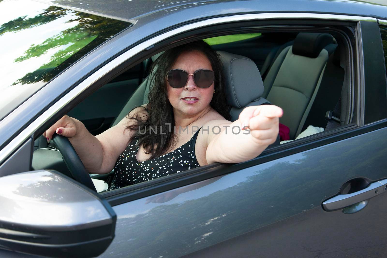 woman in sunglasses is driving and looks angry or mad pointing at you 