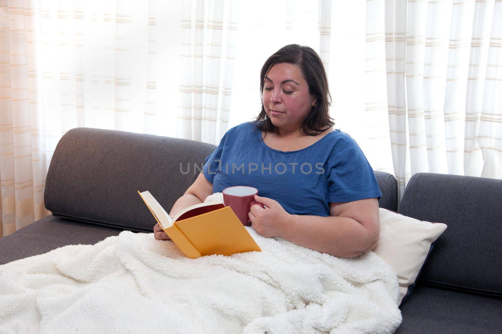 Person cuddled up on their couch with a book and mug, looking cozy and comfortable
