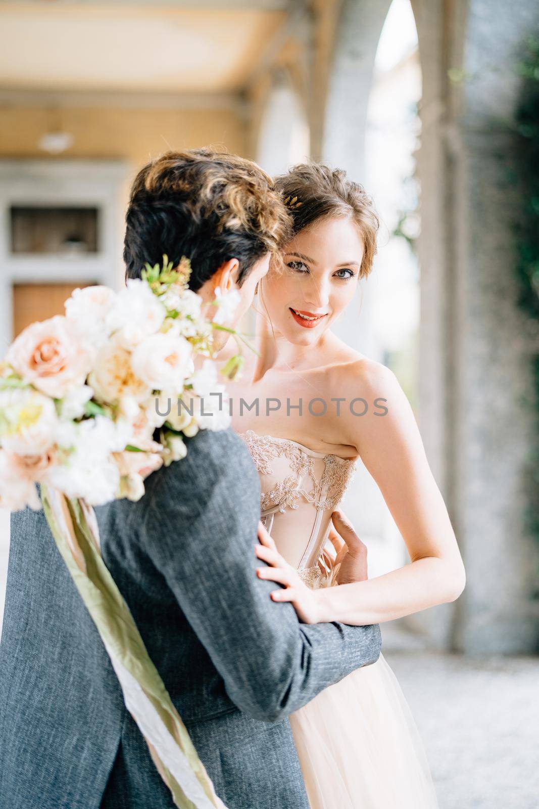 Groom hugs bride with a bouquet of flowers on an old terrace with arches entwined with green ivy. Lake Como. Close up by Nadtochiy