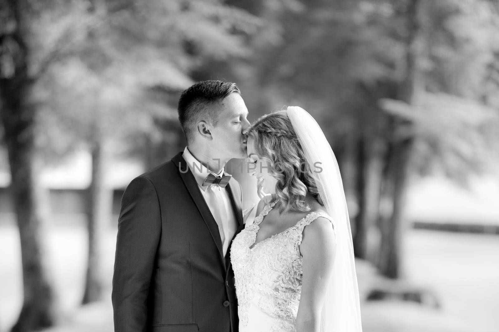 Groom hugging bride tenderly and kisses her on forehead during photo shooting in park. by kasto